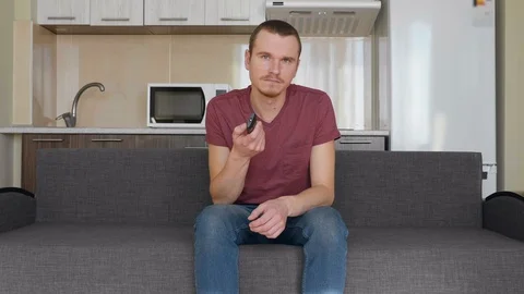 Unhappy young guy sitting on the couch and switches channels on the TV Stock Footage 107654470