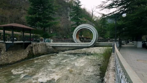 Unique modern circular bridge arching over a fast-flowing river amidst trees in Vídeo Stock 335465010