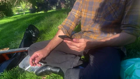 Unknown Man, Cigarette in Hand, Engrossed in Phone on Grass. Stealthy Puff Vídeos de archivo 251714579