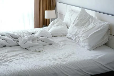 Unmade bed with crumpled white sheets and pillows, soft natural light filtering Stock Photos