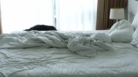 Unmade bed with rumpled white sheets and pillows, natural light streaming 스톡 동영상 330165256