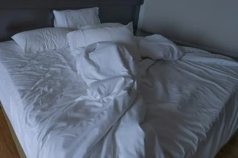 Unmade hotel bed with rumpled white sheets, pillows and duvet in dim guest .. 스톡 사진