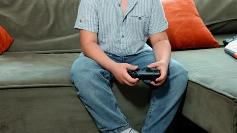 Unrecognizable boy holds game controller on sofa, striped shirt and jeans 動画素材 311506551
