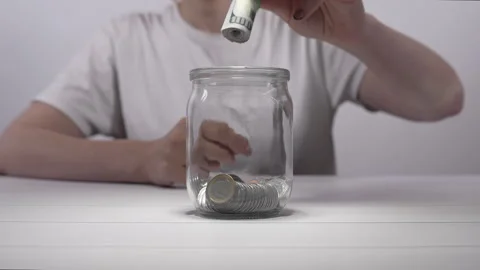 An unrecognizable girl puts a bundle of banknotes into a glass jar with coins. Stock Footage 275681599