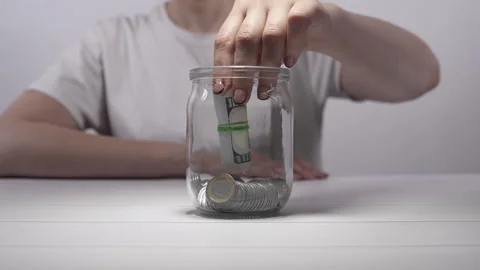 An unrecognizable girl puts a bundle of banknotes into a glass jar with coins. Stock Footage 276182797