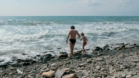 Unrecognizable man and boy go to swim in sea waves in storm on stone beach. Stock Footage 116022892