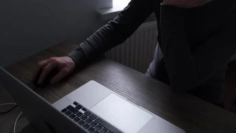 An unrecognizable man in gray sits at a table using a laptop and controls the Stock Footage 273858905