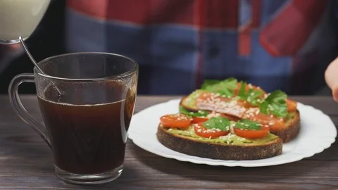 Unrecognizable man is having breakfast. Pours milk foam in coffee, two avocado Stock Footage 104769767