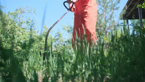 An unrecognizable man in an orange boilersuit is cutting grass with a motor trim Stock Footage 194732691