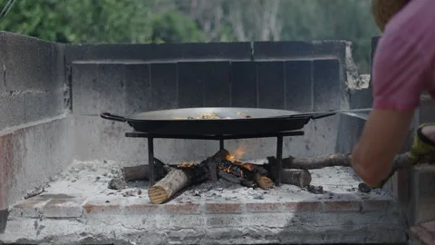 An unrecognizable man in a pink Tshirt controls the paella fire Stock Footage 259956922