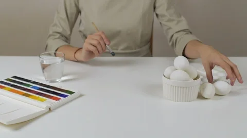 Unrecognizable woman paints Easter eggs with a brush. On the table is a glass of Stock Footage 86986582