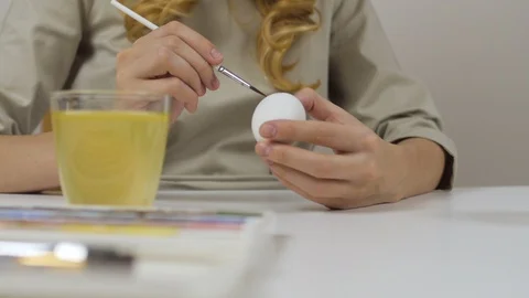 Unrecognizable woman paints Easter eggs with a brush. On the table is a glass of Stock Footage 86992148