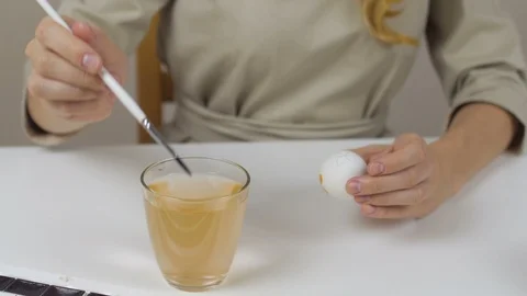 Unrecognizable woman paints Easter eggs with a brush. On the table is a glass of Stock Footage 86992885