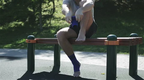An unshaved man sitting on a bench and putting on an ankle support Stock Footage 67626577