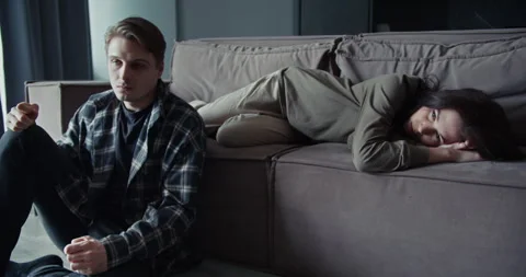 Upset angry guy sits near the sofa on which his girlfriend lies Stock Footage 305780243