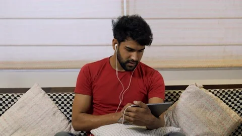 Upset Asian man using digital tablet while sitting on couch at home. Vidéo 155576486