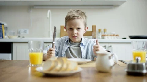 Upset boy sitting at the table with knife and fork and waiting for a dinner Stock Footage 107822961