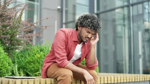 Upset businessman suffering from a headache while sitting on a bench on street  Stock Footage 302473023