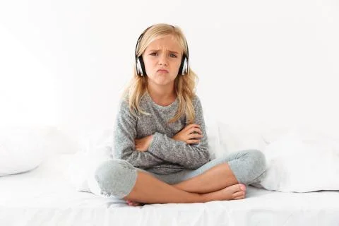 Upset female kid in headphones sitting with crossed arms and legs in bed, loo Stock Photos