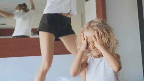 Upset little child with hyperactive mother jumping on bed on the background Stock Footage 89789885