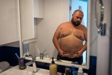 Upset man has extra weight looking at mirror touching big abdomen in bathroom at Stock Photos