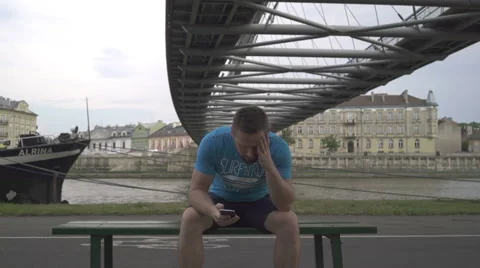 Upset man receives message, sitting one the bench against the river. 库存影片 65187710