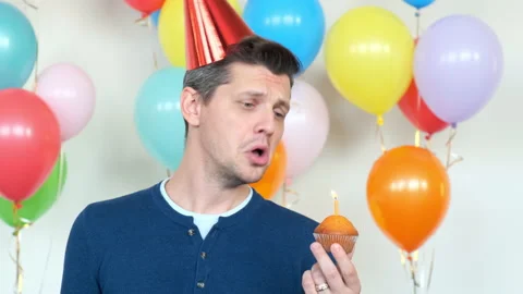 Upset man in red party cone blows burning candle on cupcake 스톡 동영상 137572553