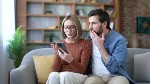 Upset married couple reading bad news on mobile phone while sitting on sofa  Stock Footage 323806608