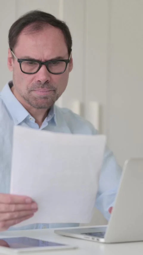 Upset Middle Aged Man Reading Documents while using Laptop, Vertical Video 스톡 동영상 339584829