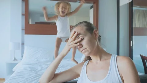 Upset mother with hyperactive child jumping on bed on the background Stock Footage 90239007