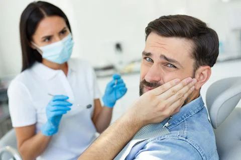 Upset patient touching face while having toothache near dentist Stock Photos