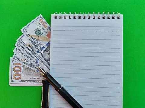 US Dollar behind lined notebook with black pen on bright green background Stock Photos