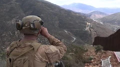 U.S. service member uses binoculars to survey the border wall from a hilltop Stock Footage 334376700