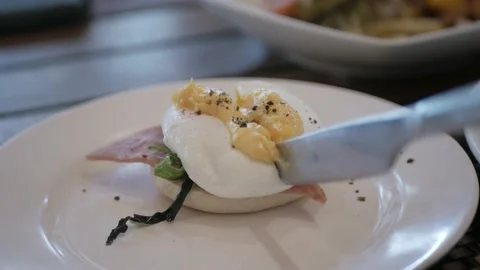 Using knife and fork cutting egg yolk to eat egg benedict in white plate. Cut Stock Footage 219999840
