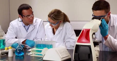 Using microscope and test tubes, scientists wearing lab coats, gloves jotting Stock Photos