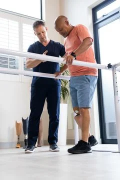Using parallel bars, man with prosthetic leg walking with therapist in rehab Stock Photos