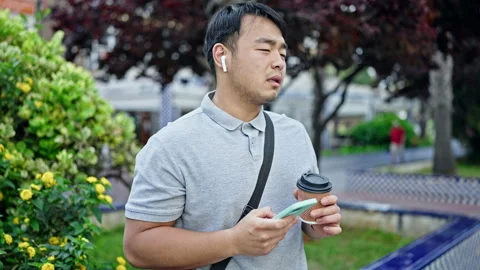 Using smartphone and earphones drinking coffee at park Stock Footage 247386760