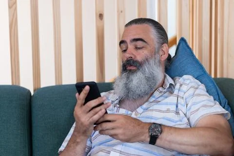 Using smartphone, bearded senior man relaxing on couch, checking messages Foto stock