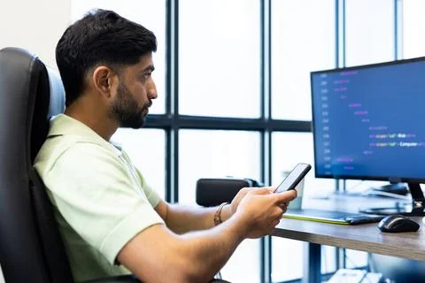 Using smartphone, Indian man sitting at desk with computer screen displaying Stock Photos