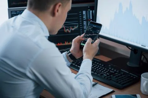 Using smartphone. Man working online in the office with multiple computer scr Stock Photos