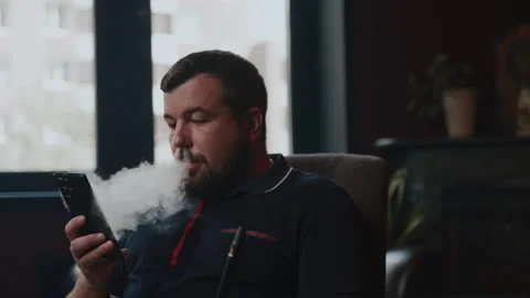 Using a smartphone while smoking. Man with beard, slow motion, bright windows on Stock Footage 253410703