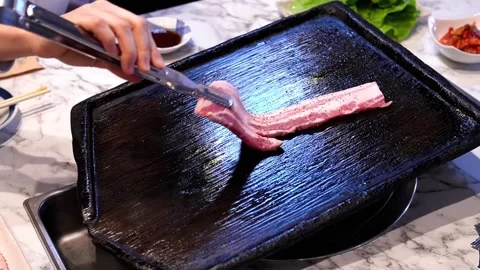 Using Tongs to Place Pork Belly on a Stone Grill in a Korean BBQ Restaurant Stock Footage 333740064