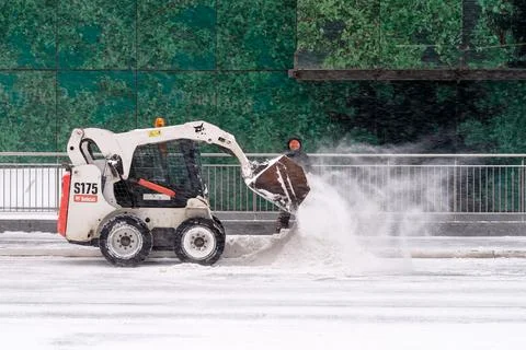 A utility worker and a small loader excavator remove snow from the road Stock Photos