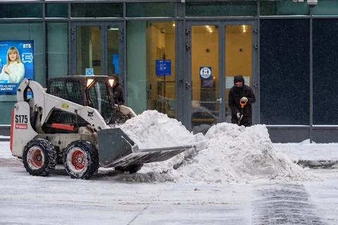 A utility worker and a small loader excavator remove snow from the road Stock Photos