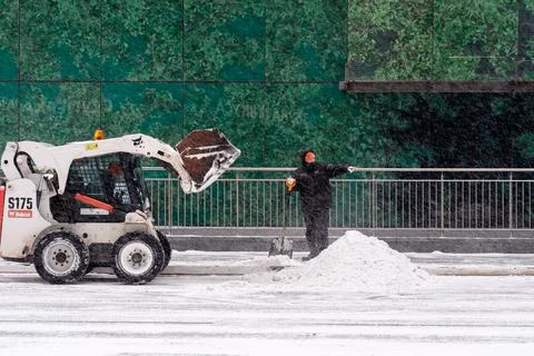 A utility worker and a small loader excavator remove snow from the road Stock Photos