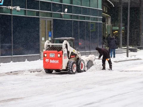 A utility worker and a small loader excavator remove snow from the road Stock Photos