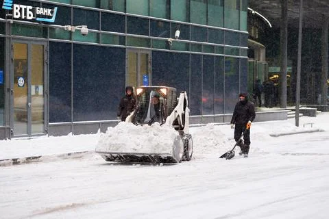 A utility worker and a small loader excavator remove snow from the road Foto stock