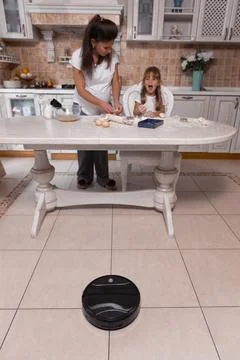 Vacuum cleaner at work in a busy kitchen while mother and daughter prepare a Stock Photos