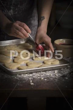 Valentine's Day baking, woman spreading raspberry jam on heart shaped biscuit Stock Photos