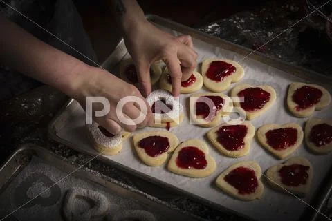 Valentine's Day baking, woman spreading raspberry jam on heart shaped biscuit Stock Photos